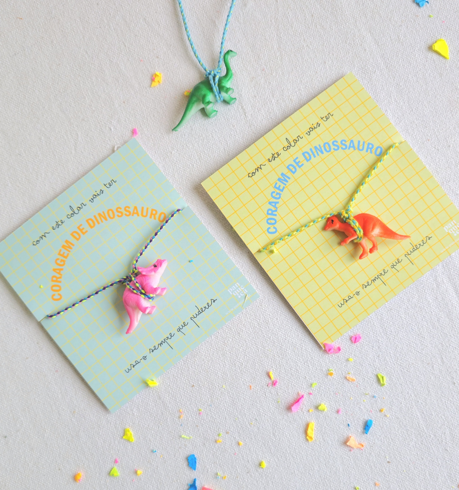 Colares infantis com pendentes de dinossauros em cartões coloridos sobre superfície clara com confete
