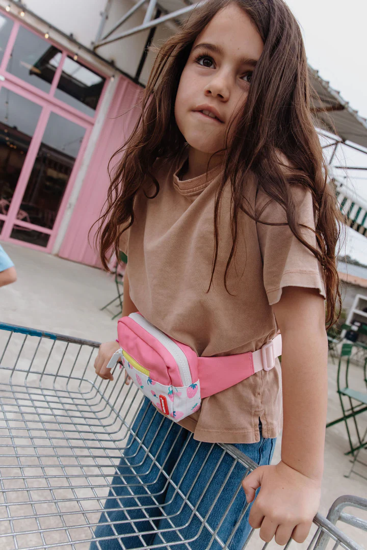 Criança com t-shirt bege e pochete rosa empurrando carrinho de compras