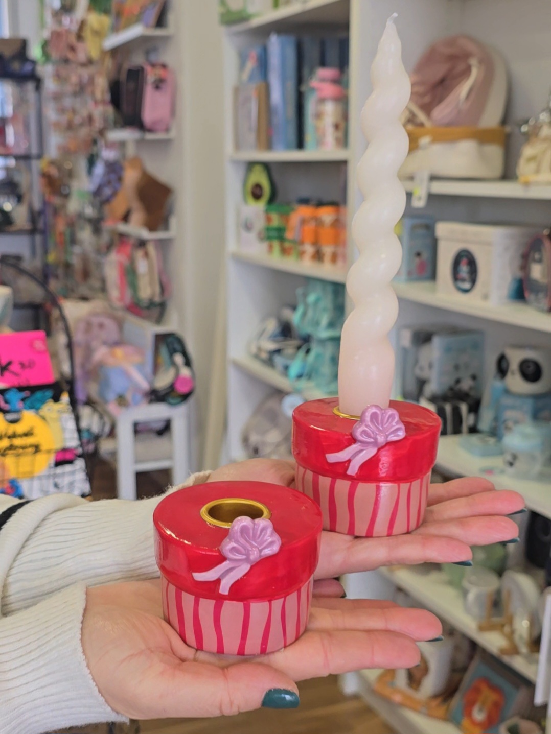 Castiçais vermelhos com vela branca torcida e decoração em fita lilás, mãos femininas, loja ao fundo.
