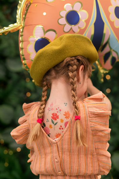 Menina com boina verde e vestido laranja com riscas e tatuagens de flores nas costas, segurando balão cor-de-rosa com padrão floral