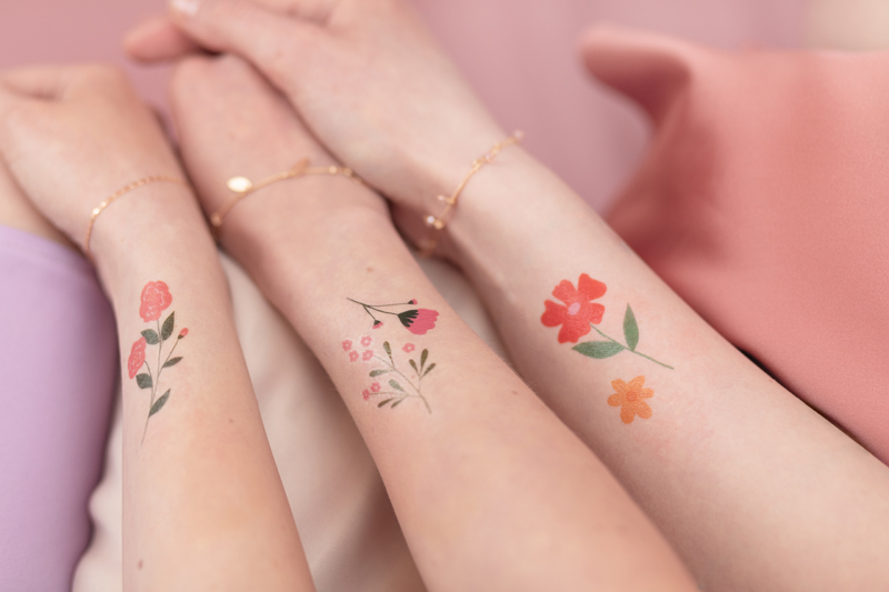 Três braços com tatuagens florais coloridas em fundo rosa e lilás
