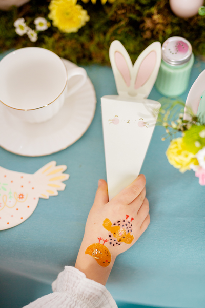 Embalagem de papel em forma de coelho branco com orelhas, mão de criança com tatuagens temporárias e chávena branca numa mesa azul