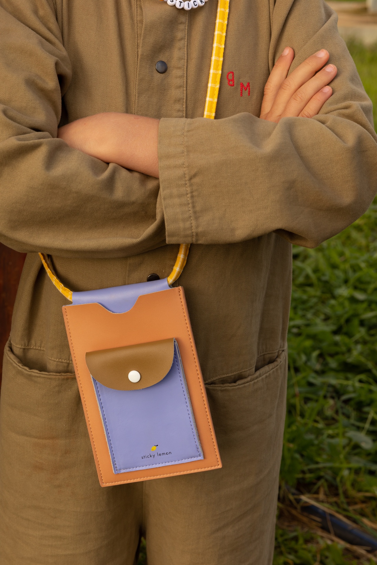 Bolsa pequena de couro azul e castanho com correia amarela xadrez usada por pessoa de macacão castanho