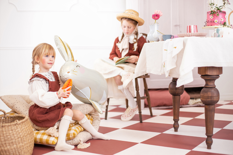 Duas crianças numa sala com chão xadrez, uma com balão coelho, outra a ler junto a mesa com bolo