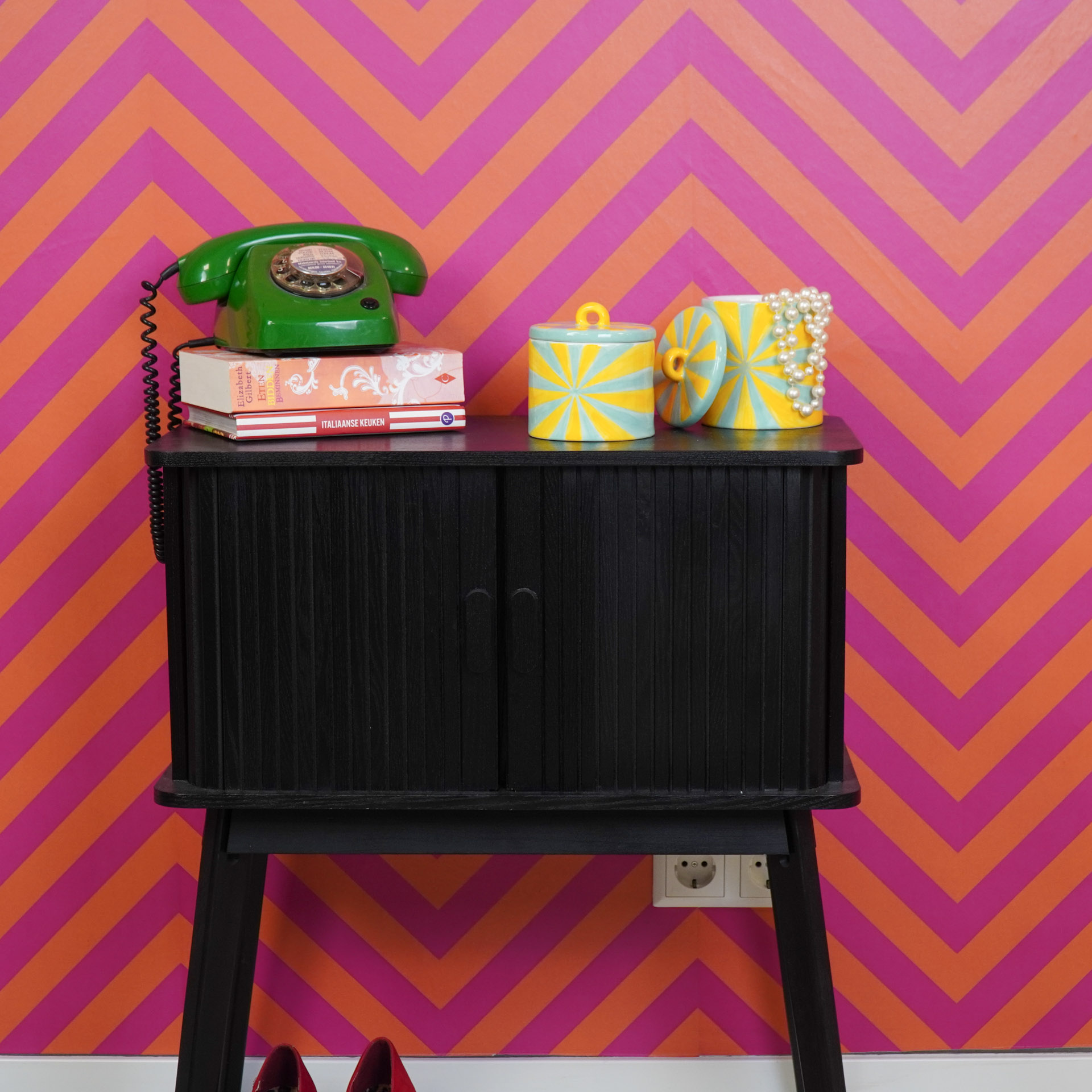 Aparador preto com telefone retro verde, livros e recipientes cerâmicos amarelos e azuis em fundo de parede rosa e laranja