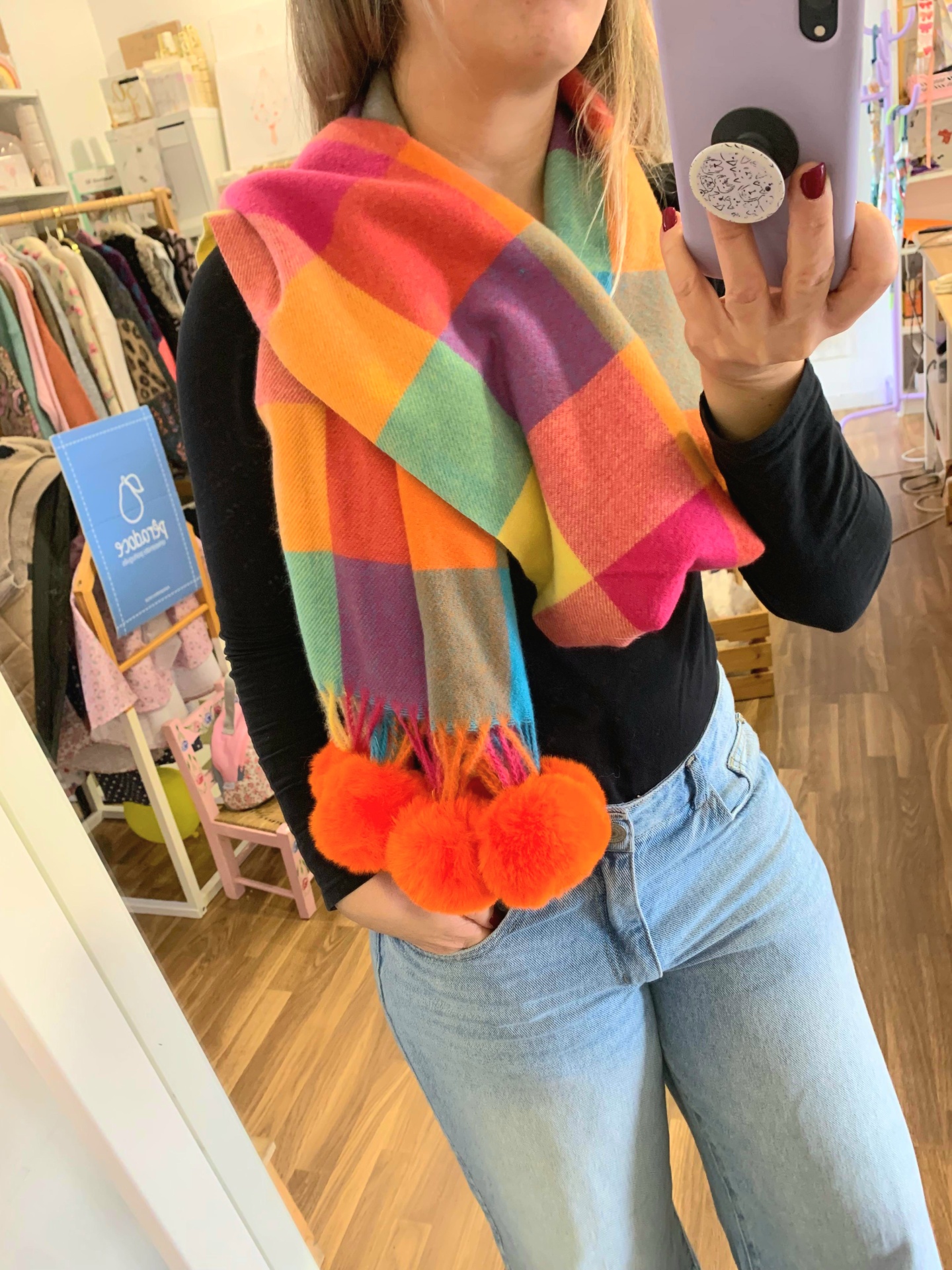 Cachecol colorido de lã com pompons laranja, usado por uma mulher com camisa preta e jeans azuis.