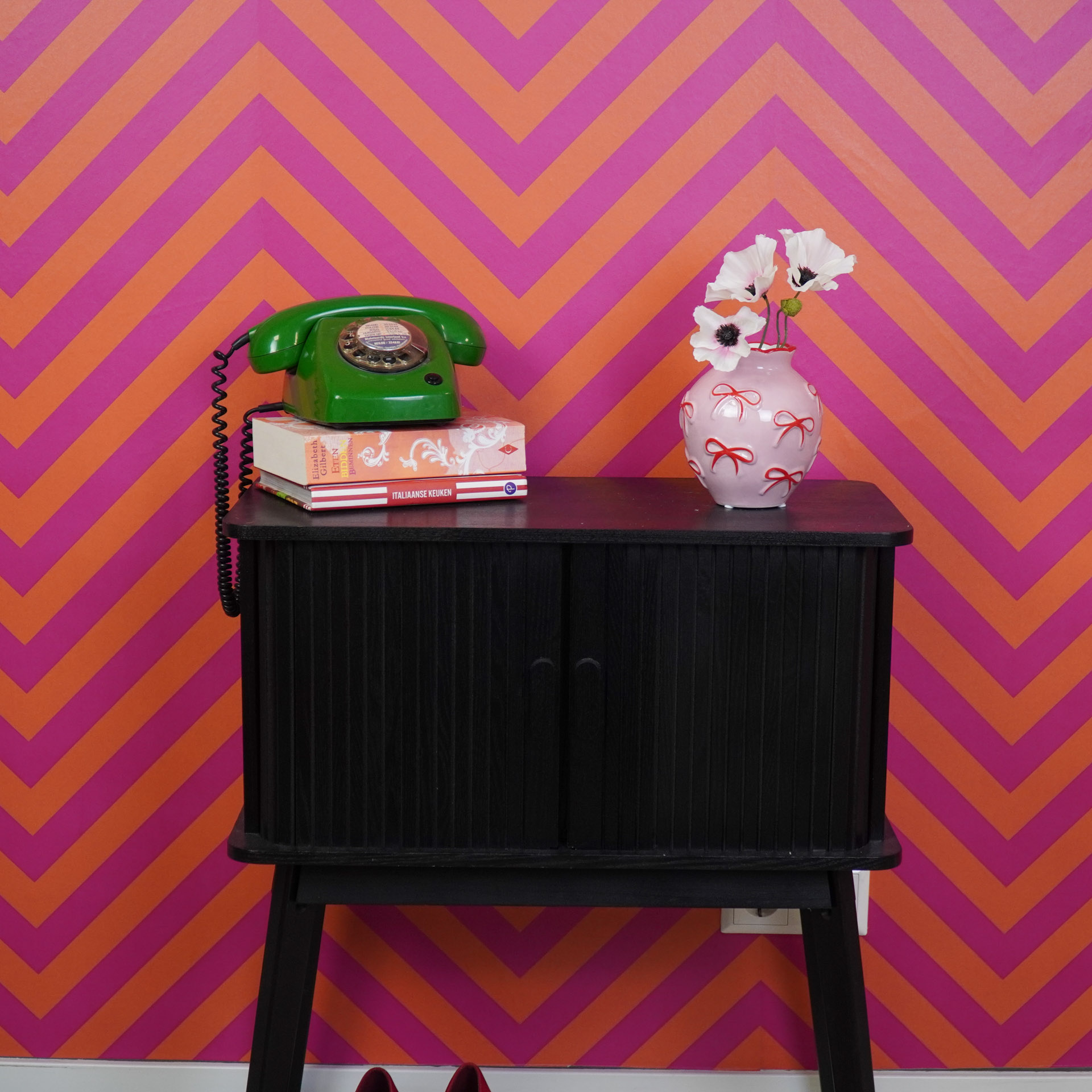 Armário preto com telefone verde vintage, livros e vaso rosa com flores contra parede laranja e rosa