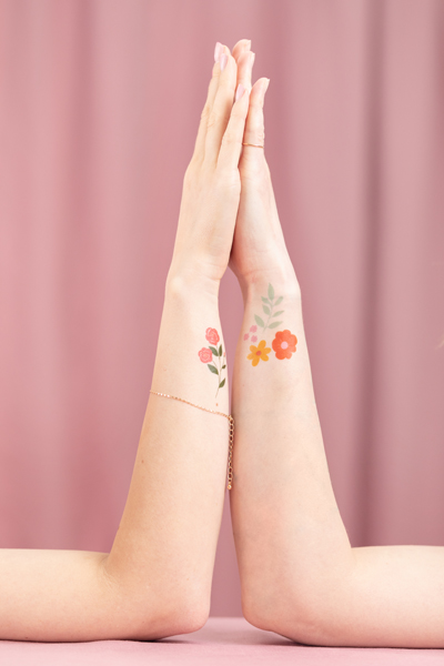 Antebraços femininos com tatuagens de flores, pulseira e anel num fundo rosa.