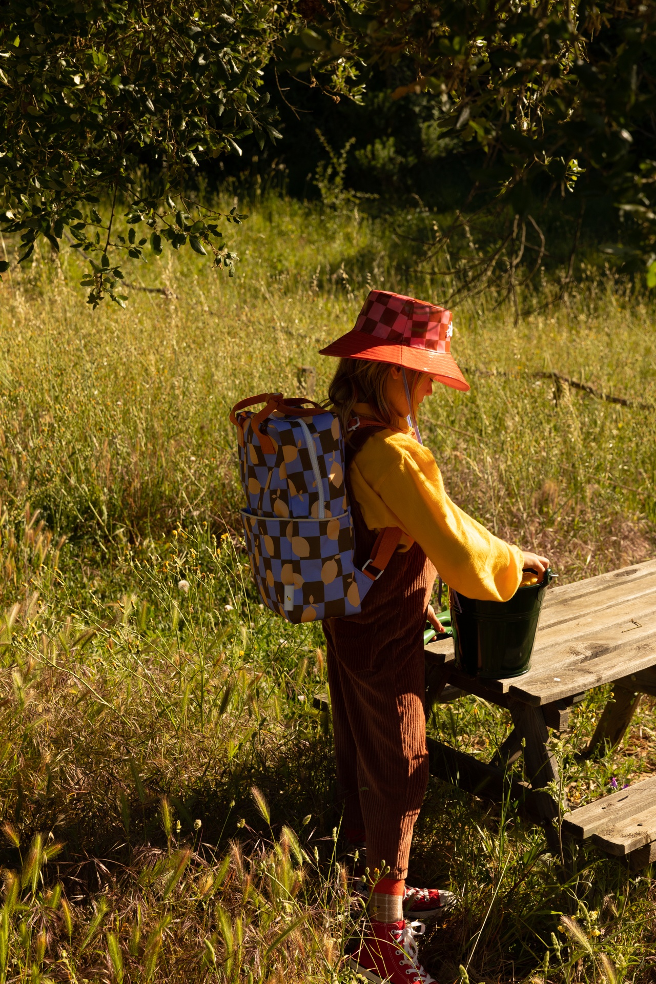 Criança com mochila colorida e chapéu vermelho junto a banco de madeira em campo verde