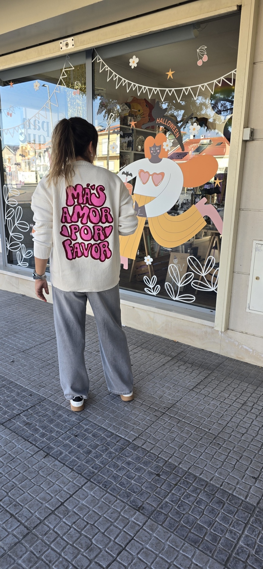 Pessoa de costas com camisola branca de malha com texto rosa, calças cinzentas e tênis brancos em frente a montra decorada