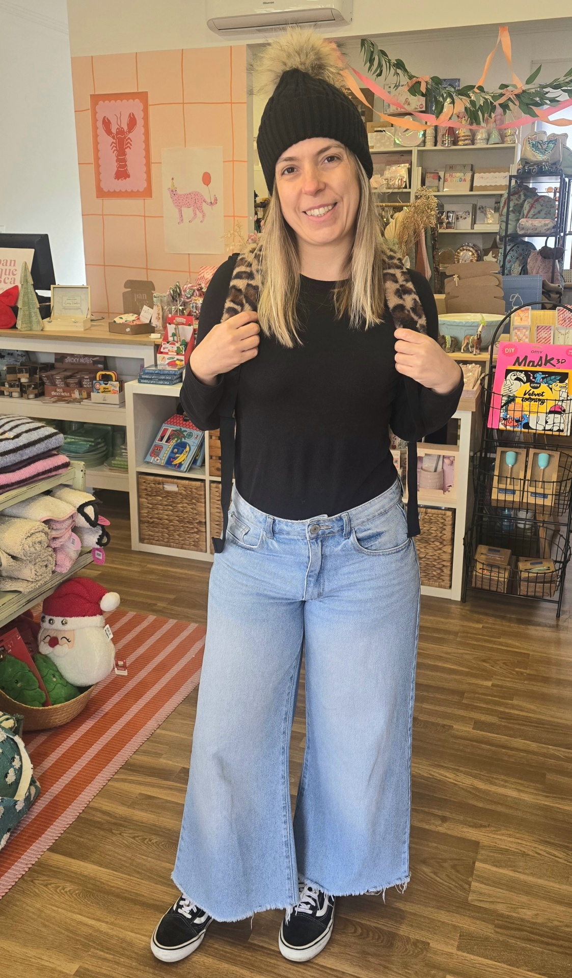 Mulher a usar jeans largos azul claro, camisola preta, boné com pompom e mochila animal print numa loja