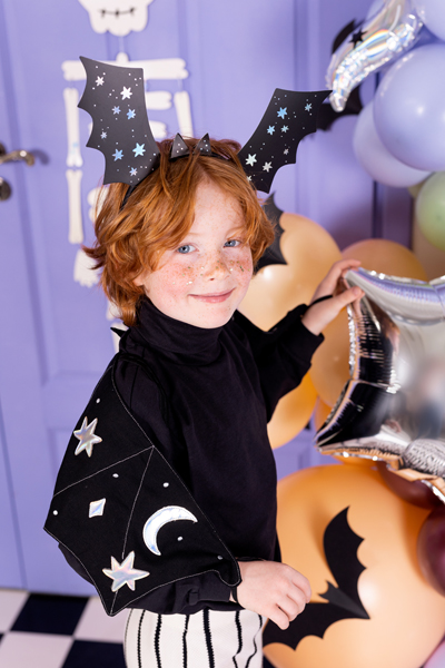 Criança com fantasia de morcego segurando balão prateado em festa colorida.