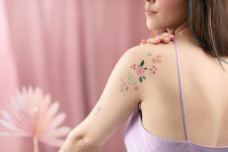 Mulher com tatuagens florais no ombro usando vestido lilás em fundo rosa