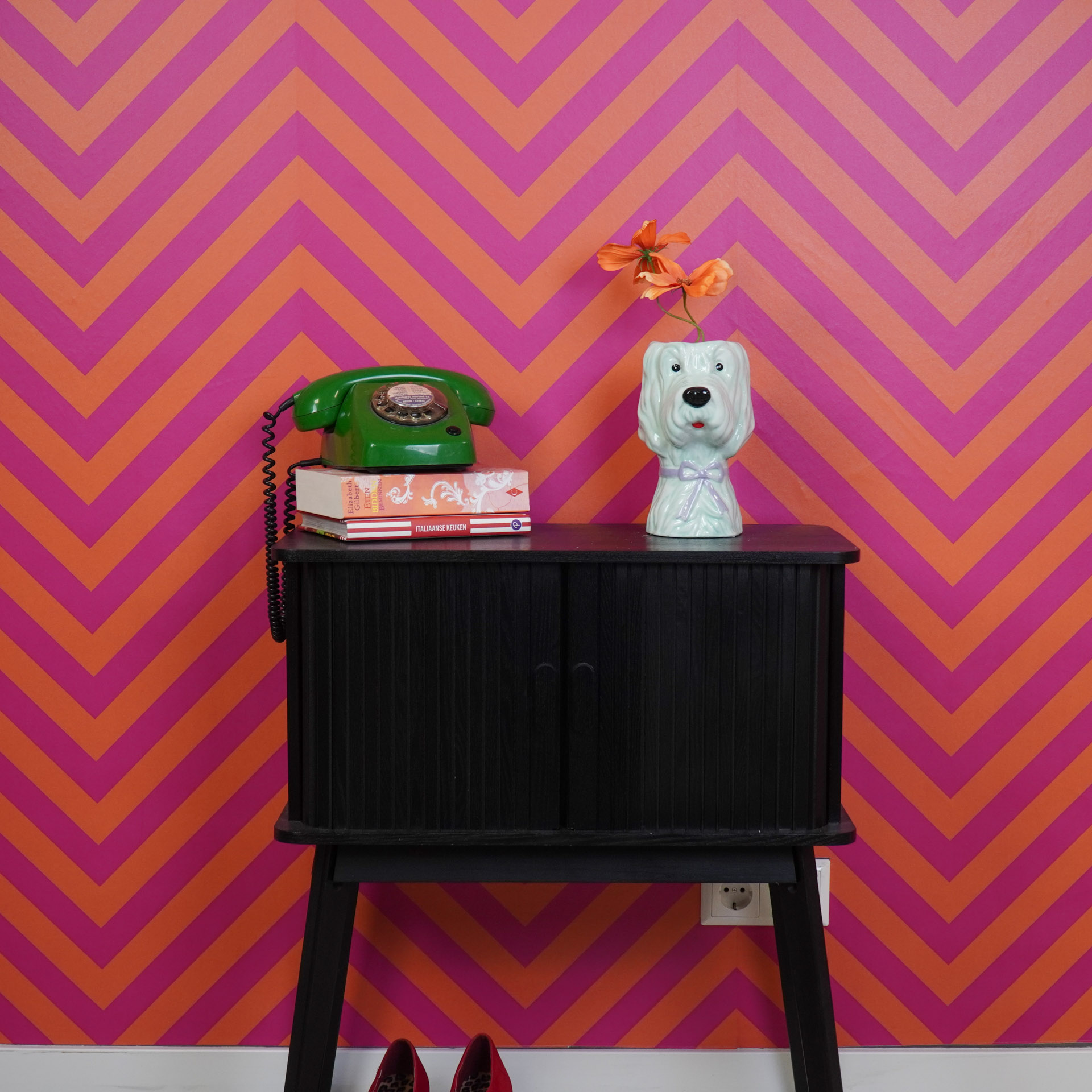 Móvel preto com telefone verde, livros e vaso de cão com flores, fundo papel de parede ziguezague laranja e rosa