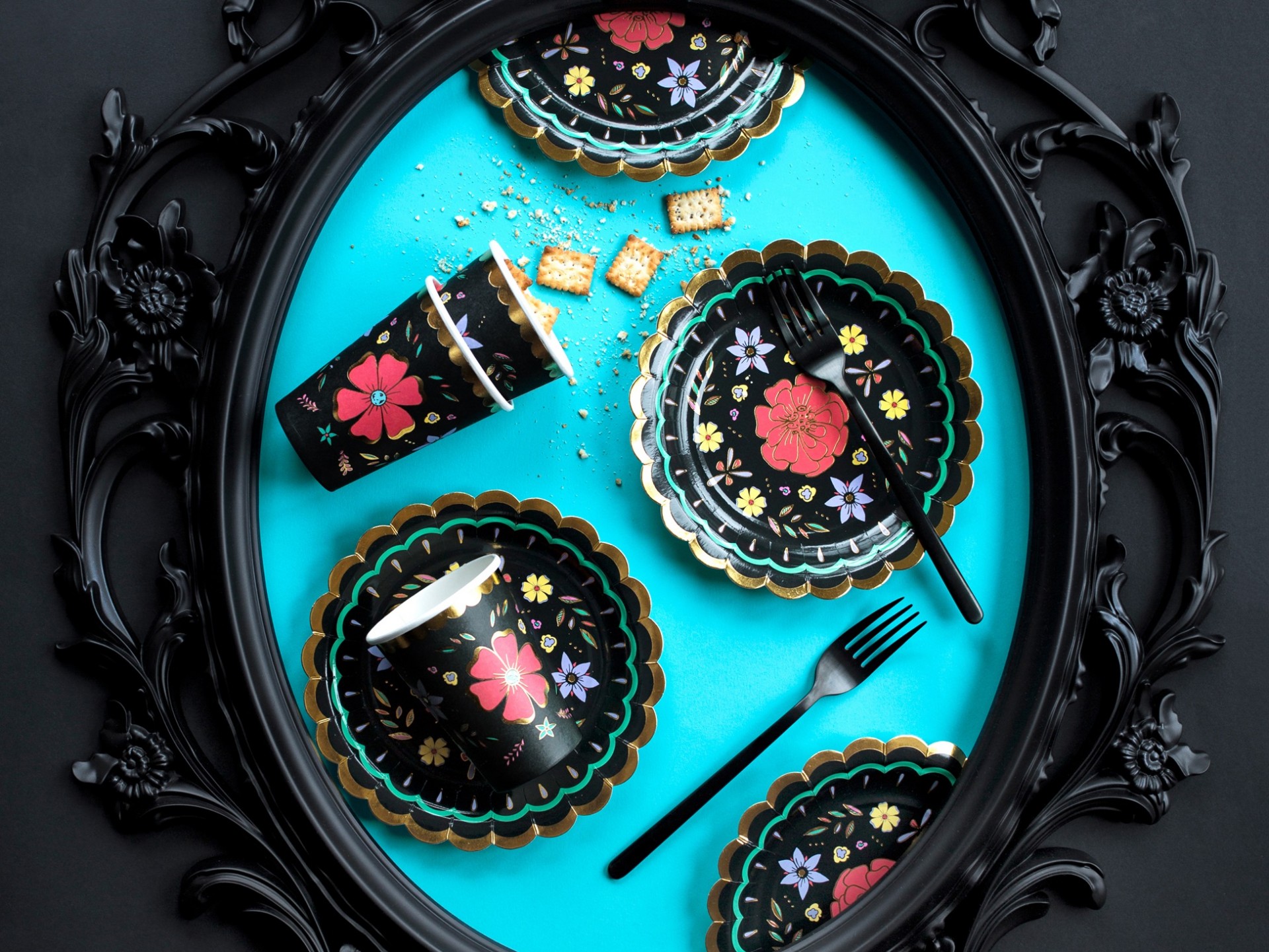 Louça de papel preta com flores coloridas e talheres pretos numa moldura de quadro com fundo azul