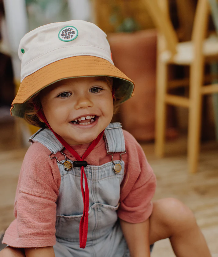 Criança com chapéu branco e castanho, camiseta rosa e jardineira de ganga sentada