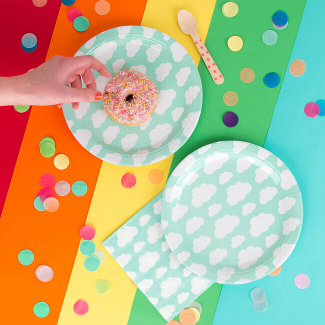 Dois pratos e guardanapo com padrão de nuvens, donut rosa com granulados, espátula de madeira, fundo colorido com confetes