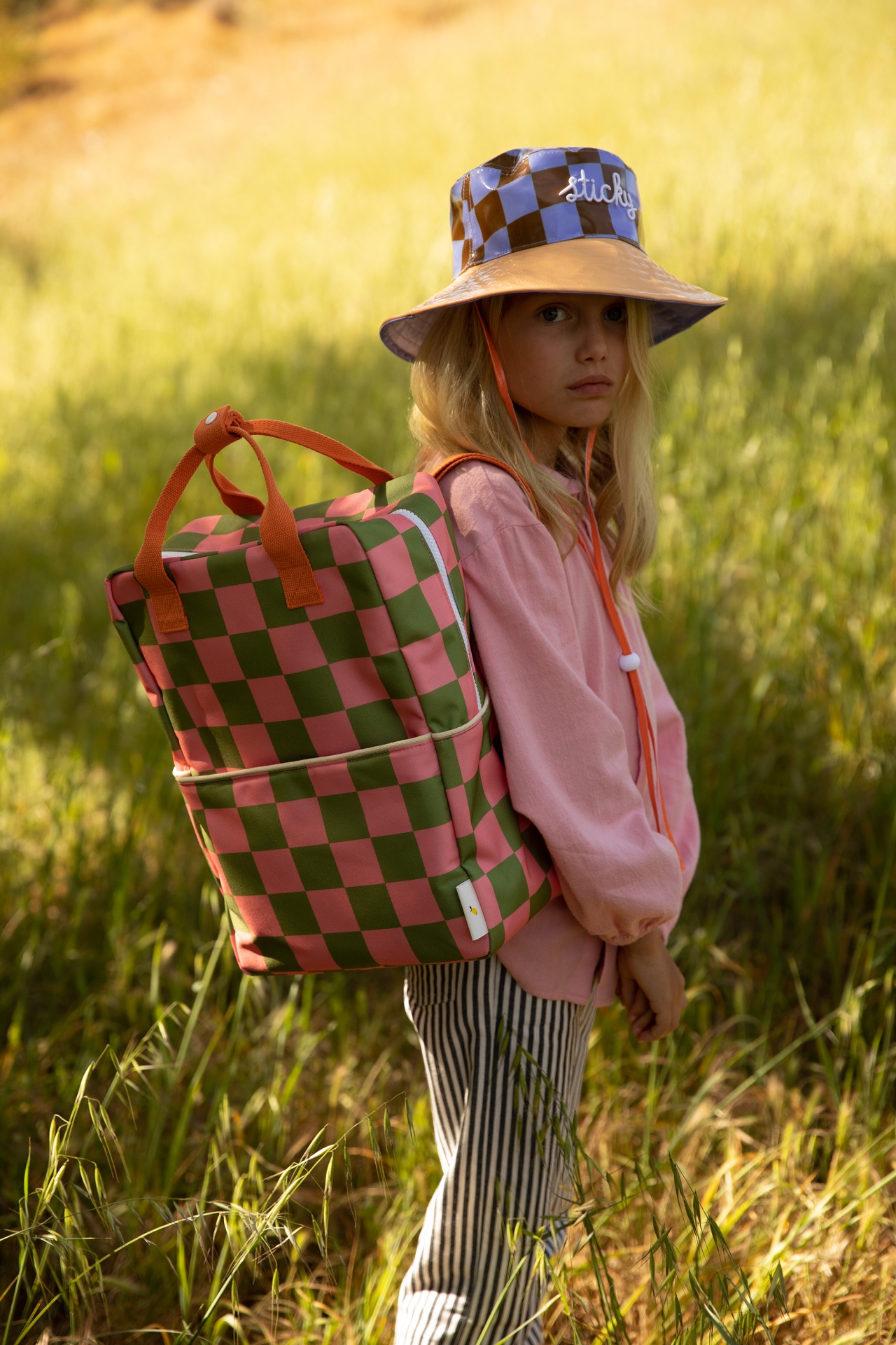 Criança com mochila xadrez rosa e verde e chapéu bucket xadrez azul e lilás em campo de relva