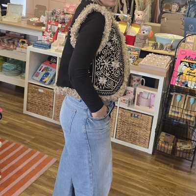 Mulher com colete bordado e saia de ganga dentro de loja com objetos