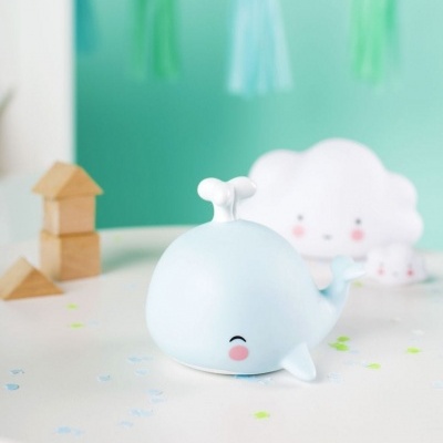 Brinquedo baleia azul claro com rosto sorridente sobre superfície branca com confetes em fundo verde.