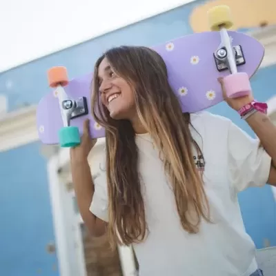 Jovem segurando prancha de skate lilás com flores e rodízios coloridos sobre os ombros, ao ar livre.