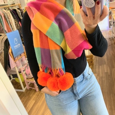 Cachecol colorido de lã com pompons laranja, usado por uma mulher com camisa preta e jeans azuis.