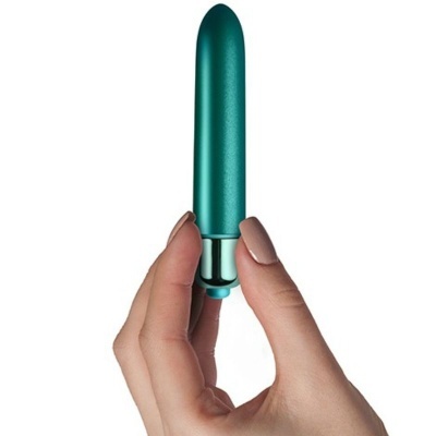 Vibrador cilíndrico verde metálico segurado por uma mão com unhas pintadas castanho claro, fundo branco.