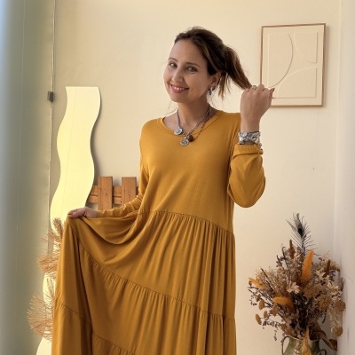 Mulher com vestido amarelo mostarda comprido com mangas longas em ambiente decorado com vaso de flores secas e espelho ondulado.