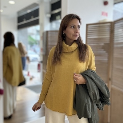 Mulher vestindo camisola amarela de gola alta e calças brancas, segurando casaco verde escuro em ambiente interior com espelho e divisória.