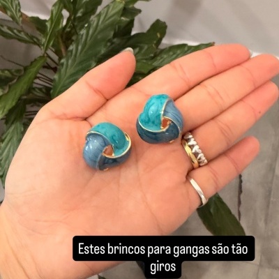 Brincos entrelaçados azul turquesa e azul claro com detalhes dourados numa mão com anéis e pulseira, usada em ambiente interior com plantas ao fundo.