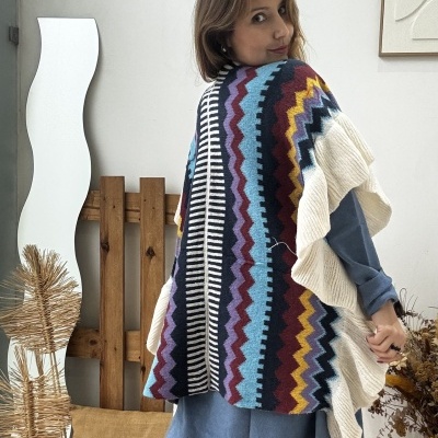 Poncho colorido em padrão zig-zag usado por mulher com vestido azul e botas pretas em ambiente interior.