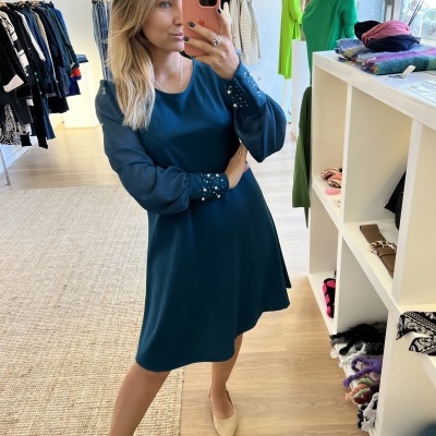 Mulher a vestir vestido azul petróleo com detalhes em pérolas nas mangas, numa loja de roupa.