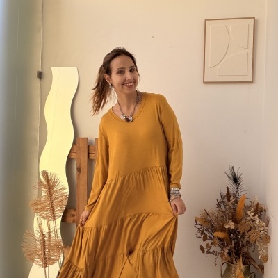Mulher com vestido amarelo mostarda comprido e botas pretas em ambiente minimalista com vaso de flores secas e espelho ondulado