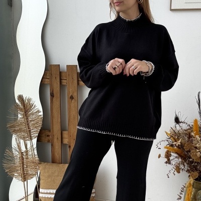 Mulher com conjunto de malha preta com detalhe branco, sapatos bege, em espaço com espelho ondulado, almofada castanha e vaso com plantas secas.