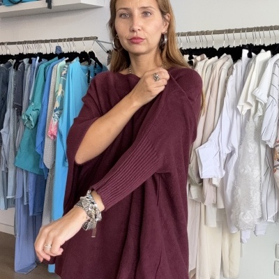 Mulher com camisola borgonha de malha e jeans numa loja de roupas com cabides ao fundo.