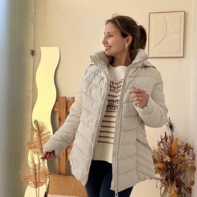 Mulher com casaco acolchoado bege claro em ambiente interior decorado com flores secas e almofada.