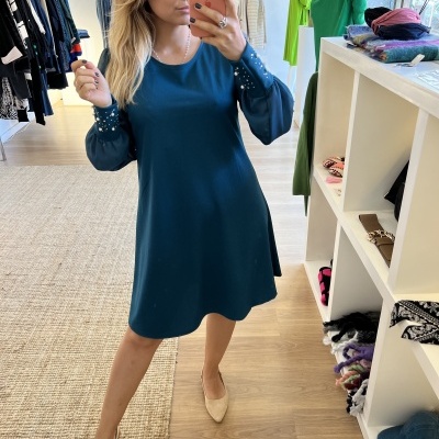 Mulher de vestido azul-escuro com detalhes de pérolas nas mangas tirando selfie numa loja de roupa