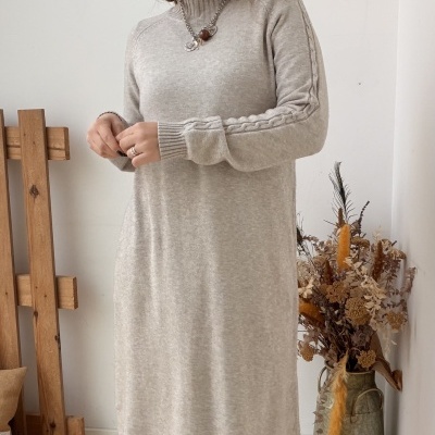 Vestido de malha bege claro com gola subida e padrão trançado nas mangas, usado com botas pretas, em ambiente com suporte de madeira, jarra com flores secas e arte na parede.