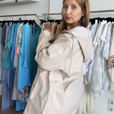 Mulher veste casaco bege claro com capuz em loja de roupa com cabides e parede branca