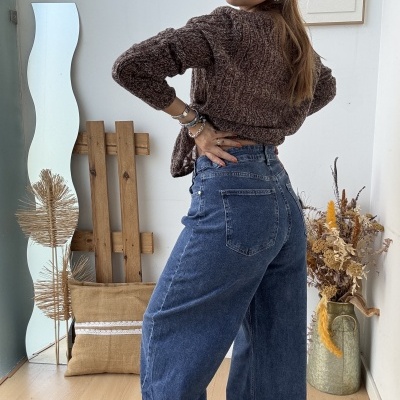 mulher com camisola castanha de malha e calças de ganga azuis largas em ambiente interior com decoração rústica