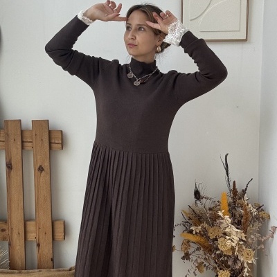 Mulher com vestido castanho de malha plissada, botins pretos, colar medalhas, em ambiente minimalista com vaso, almofada e quadro abstrato