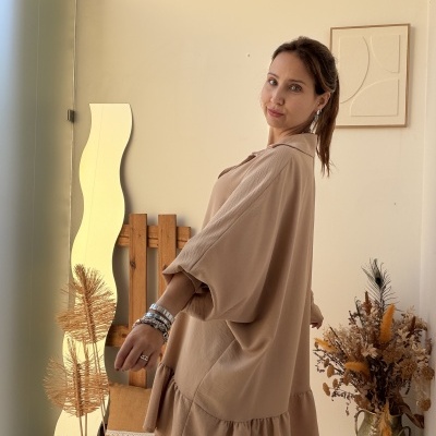 Mulher a usar túnica castanha clara com folho e pulseiras em ambiente com espelho ondulado e flores secas