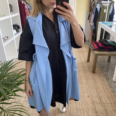 Mulher a tirar selfie com vestido preto e colete azul claro numa loja com tapete de fibra e planta verde