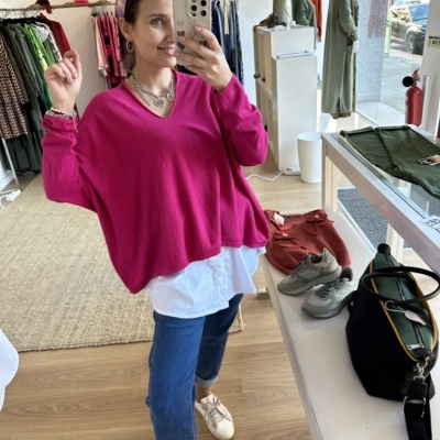 Mulher com camisola rosa, camisa branca, calças de ganga azuis e sapatilhas brancas numa loja de roupa com ténis, camisola castanha e mala preta sobre mesa branca