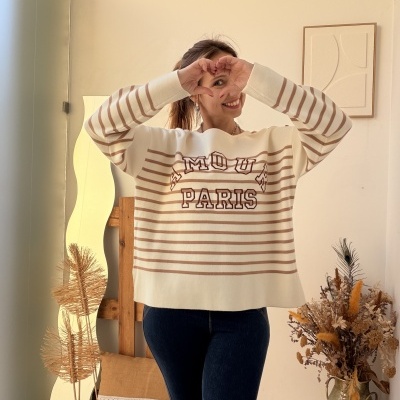 Pessoa com camisola creme às riscas castanhas e jeans escuros em sala decorada com almofada e flores secas