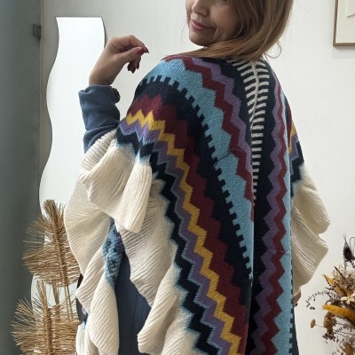 Poncho de malha colorido com padrão zigzag e franjas onduladas em ambiente interior com plantas secas e espelho