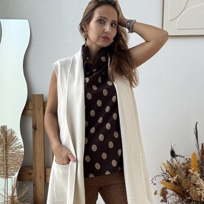 Mulher com colete branco, blusa preta com bolinhas e calças castanhas em ambiente minimalista.