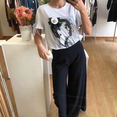 Mulher com t-shirt branca estampada e calças pretas largas num ambiente de loja de roupas.