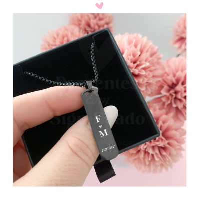 Colar pendente metálico preto gravado com letras e data, num estojo preto com flores cor-de-rosa ao fundo
