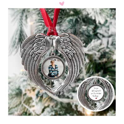 Enfeite de Natal de metal em forma de coração com asas e foto, pendurado por fita vermelha numa árvore de Natal.